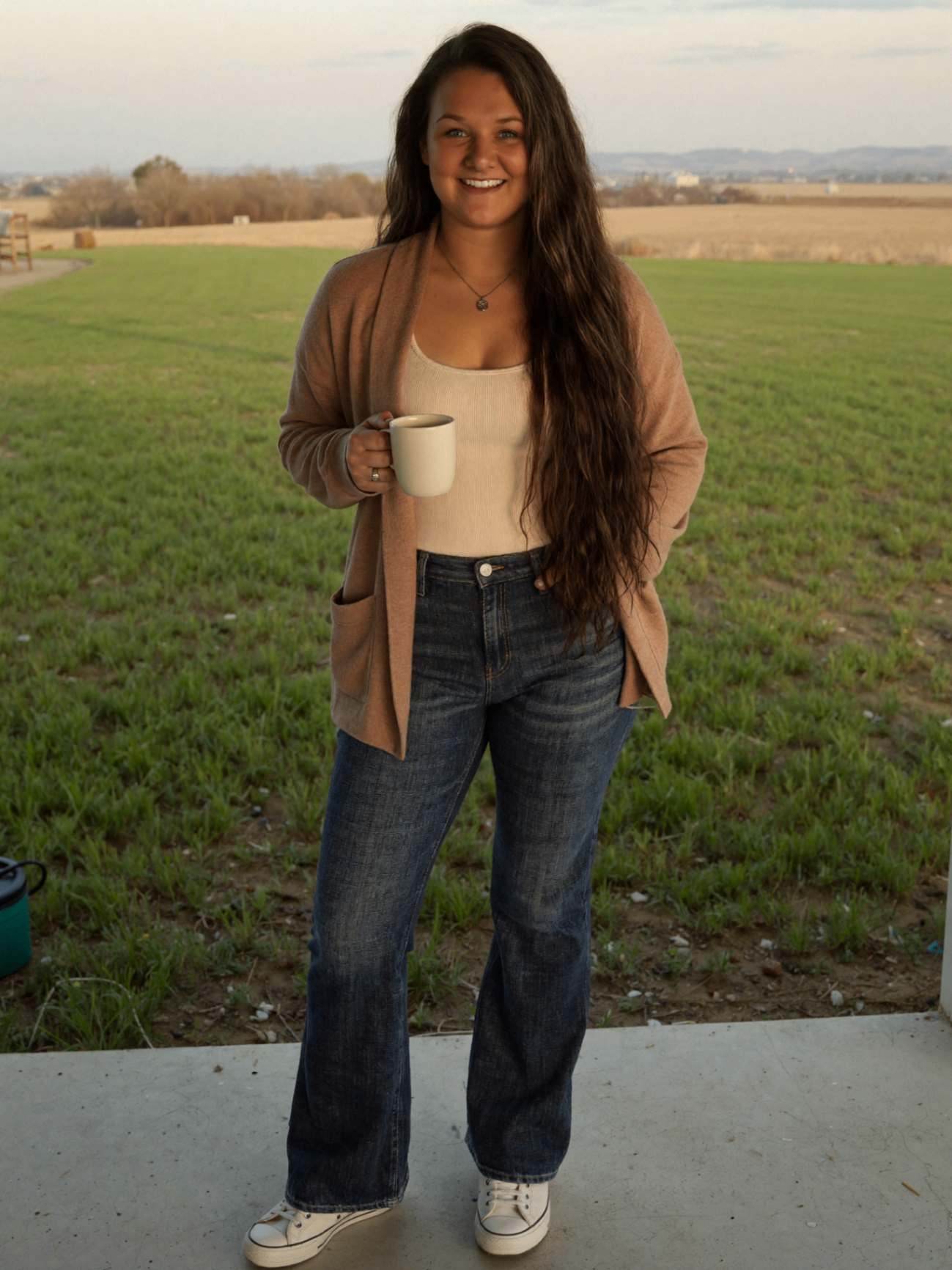 Woman holding coffee in a field, smiling warmly.