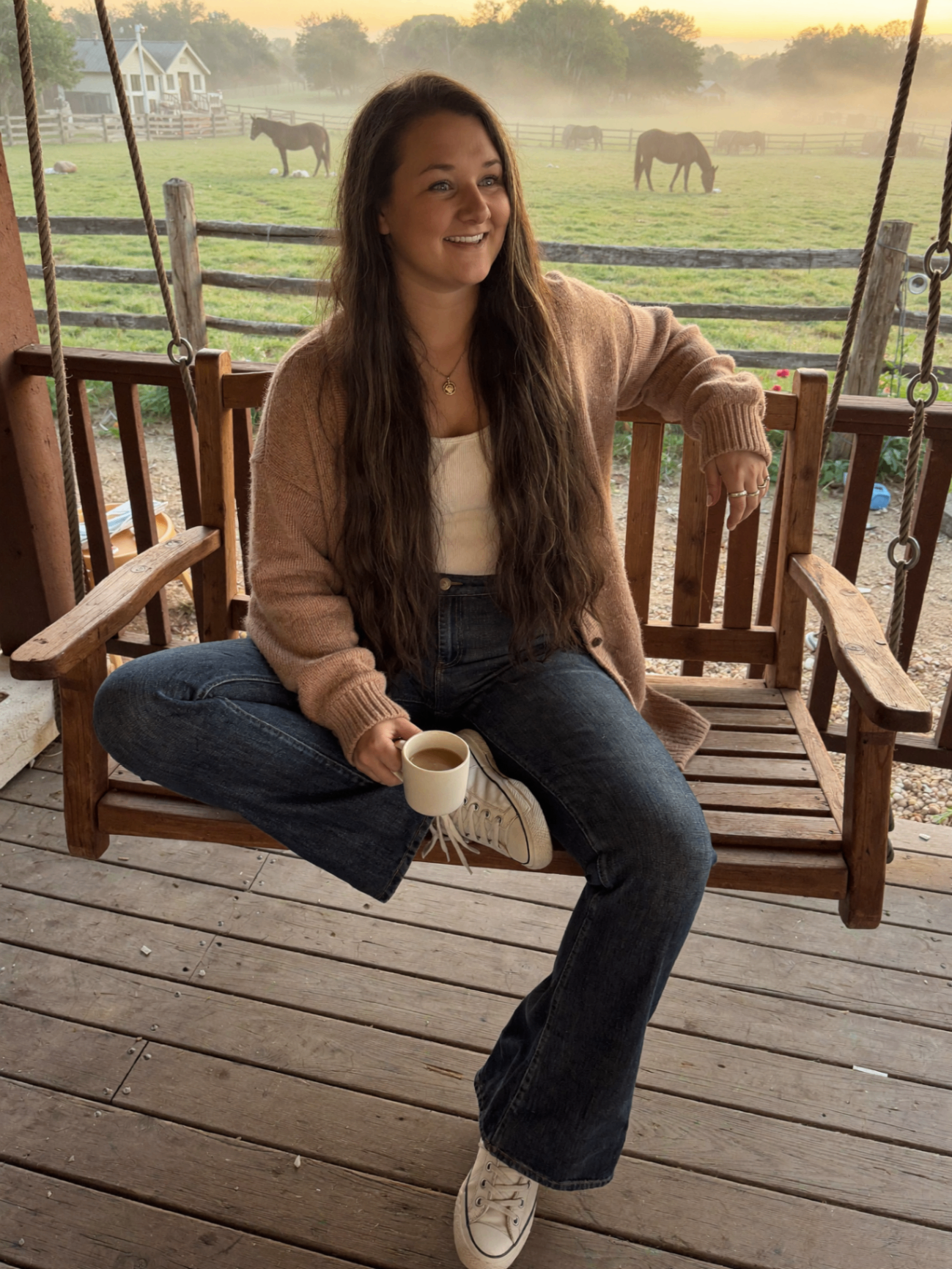 Woman on a porch at sunrise with horses in a foggy pasture.